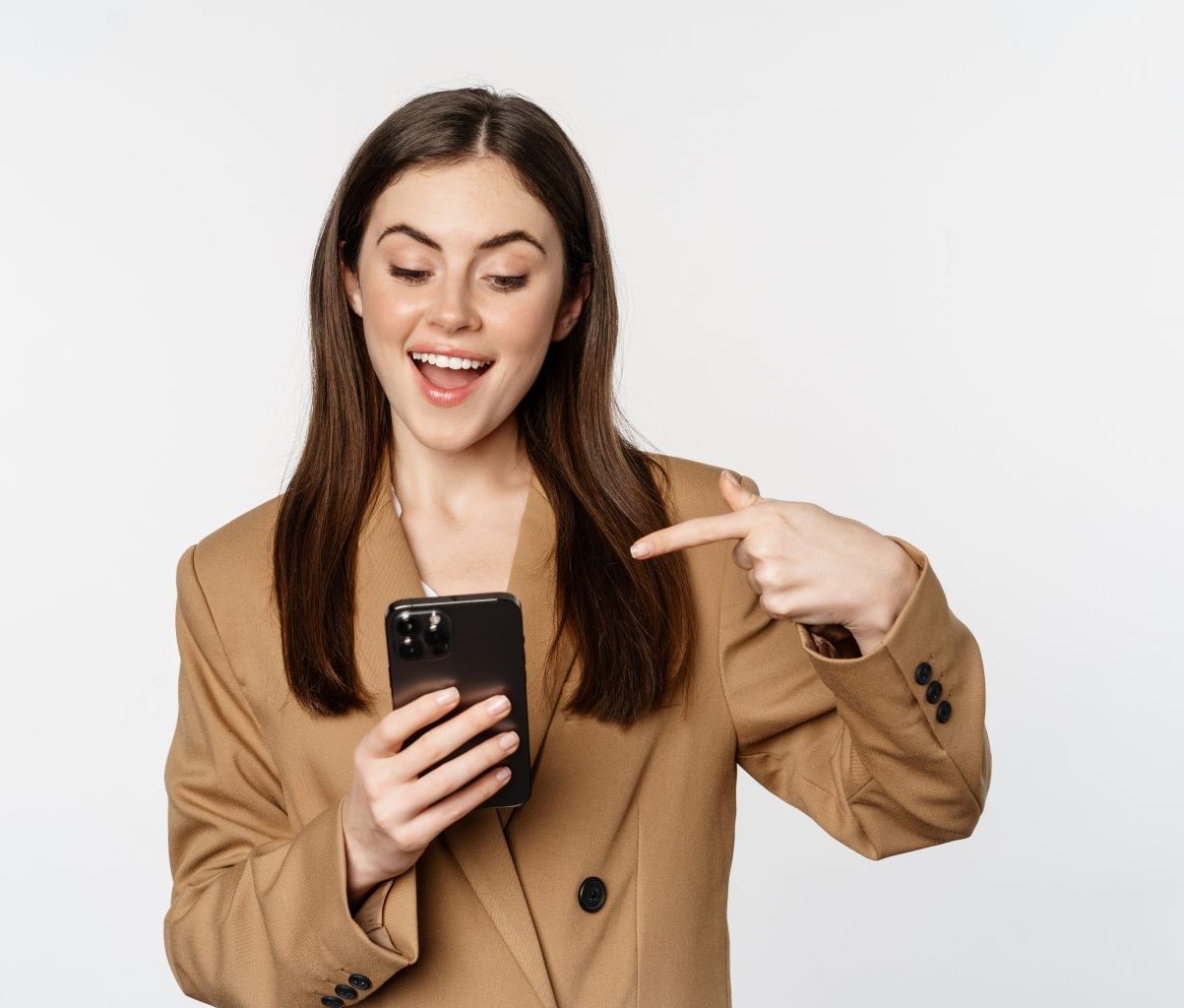 Enthusiastic saleswoman, business woman pointing finger at mobile phone and smiling, showing on cellphone, standing against white background.